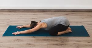 a woman is on a blue yoga mat doing child pose to relieve back pain and feel better. laying on top of her shins with the arms extended