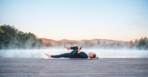 a woman is outside on a yoga mat with mountains and tree in the background. she is doing the pose half wind reliever which involves laying down and hugging one shin into her chest. this will help her relieve back pain and feel better