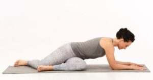 a woman is on a yoga mat doing deer pose to relieve back pain and feel better. seated, legs are bent and she's leaning over them with the forearms on the floor